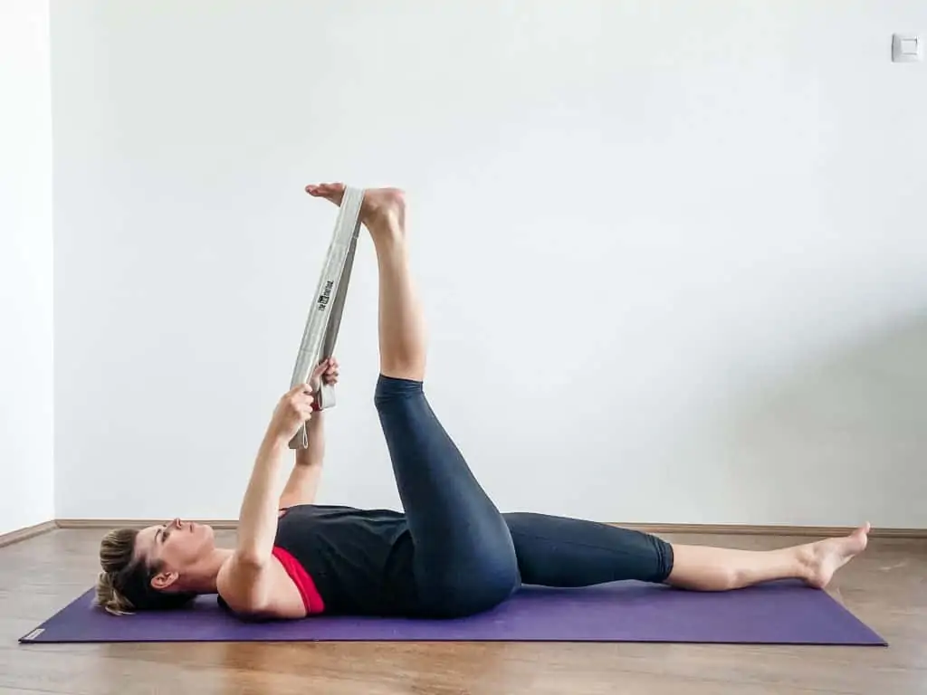 woman on a yoga mat demonstrating a supine hamstring stretch using a strap to assit