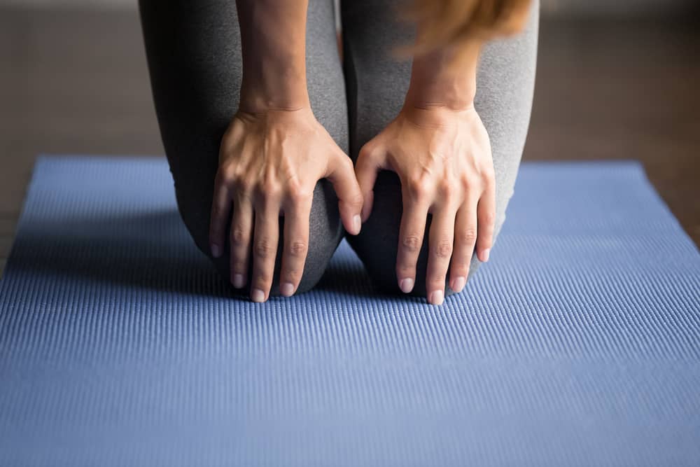 woman kneeling on a yoga mat holding her knees. Decorative image for an article about the best yoga knee pads. 